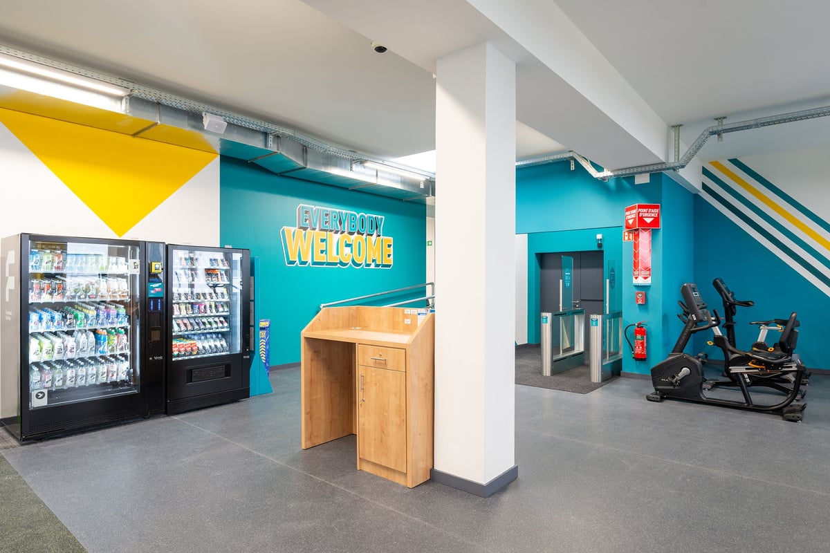 PureGym Vevey: Vélos d'exercice, distributeurs automatiques, réception dans une chambre bleu sarcelle et jaune avec un sol en bois.