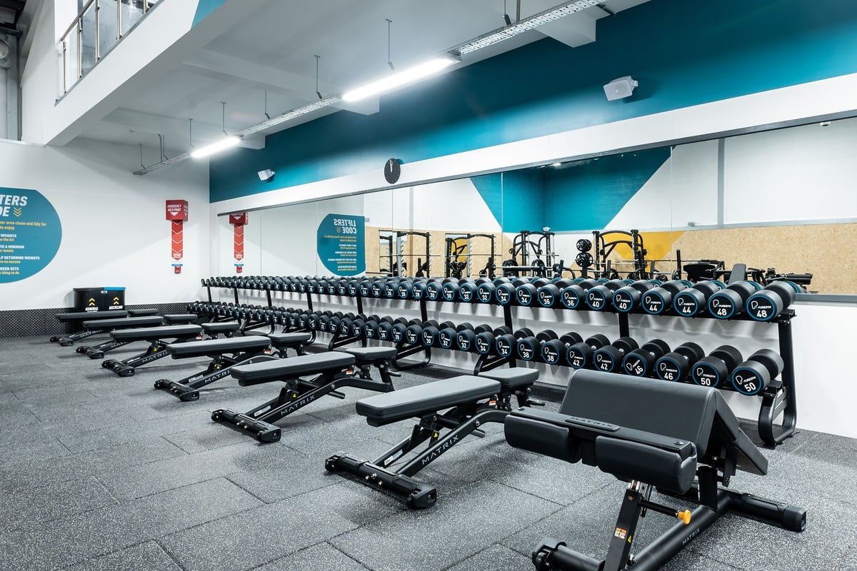 PureGym Ballymena: Rows of adjustable weight benches, dumbbells, and free weight racks on rubberized flooring.