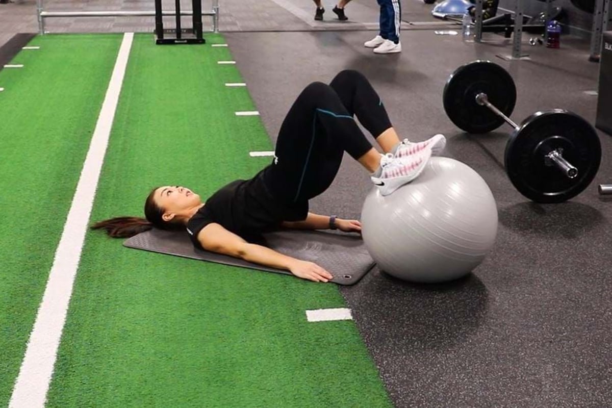 Woman exercising with legs on a stability ball on a gym mat near weights.