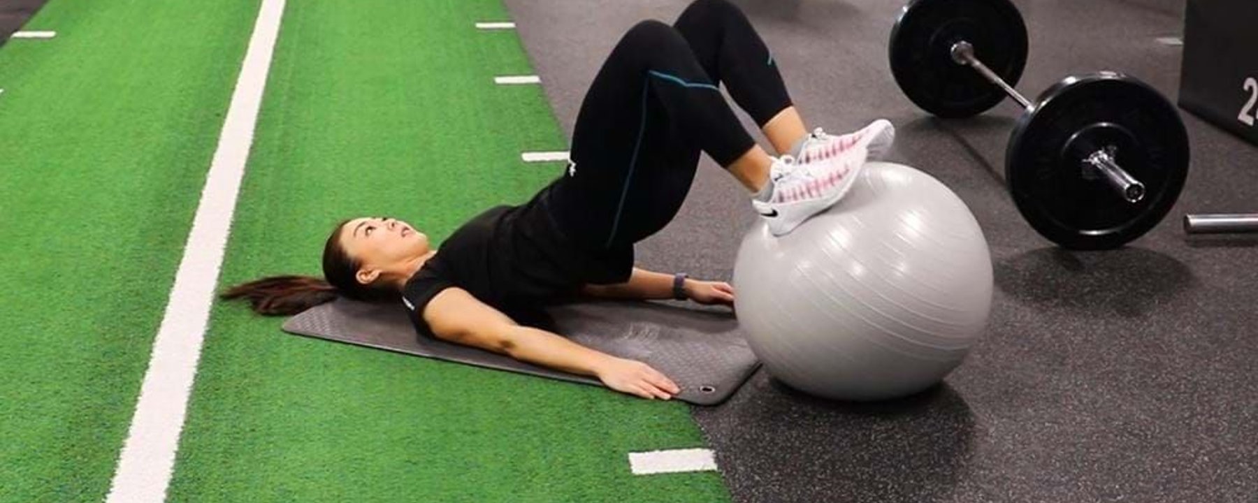 Woman exercising with legs on a stability ball on a gym mat near weights.
