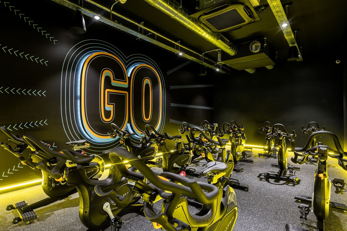 PureGym London Catford Rushey Green: Rows of spin bikes in a dimly lit studio with neon 'Go' signage and industrial ceiling.