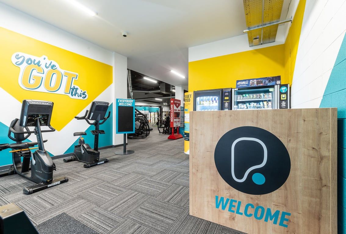 PureGym Dungannon: Exercise bikes, weights area, welcome sign, bright yellow and teal color scheme, open fitness space.