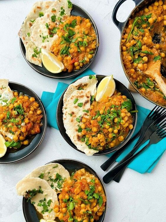 Four plates of chickpea curry with flatbread and lemon wedges next to a pan of curry