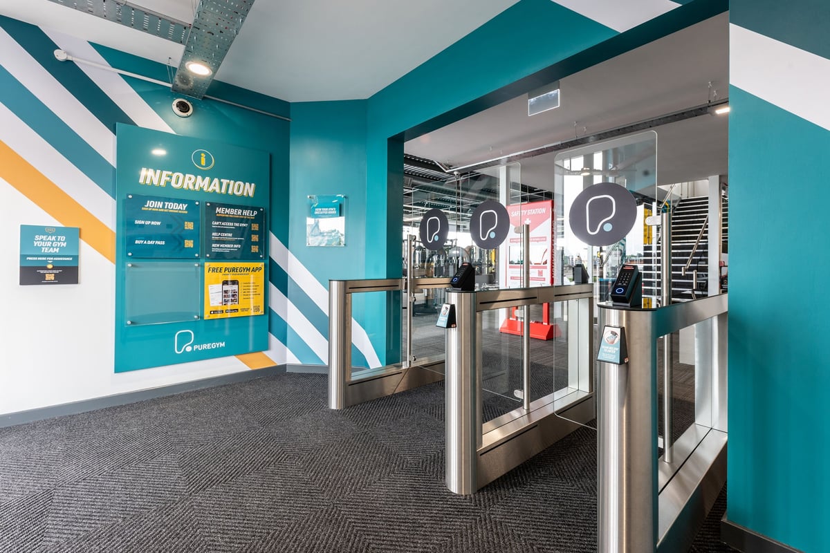 PureGym Glasgow Giffnock: Turnstiles leading into gym area, information signage, teal and white color scheme.