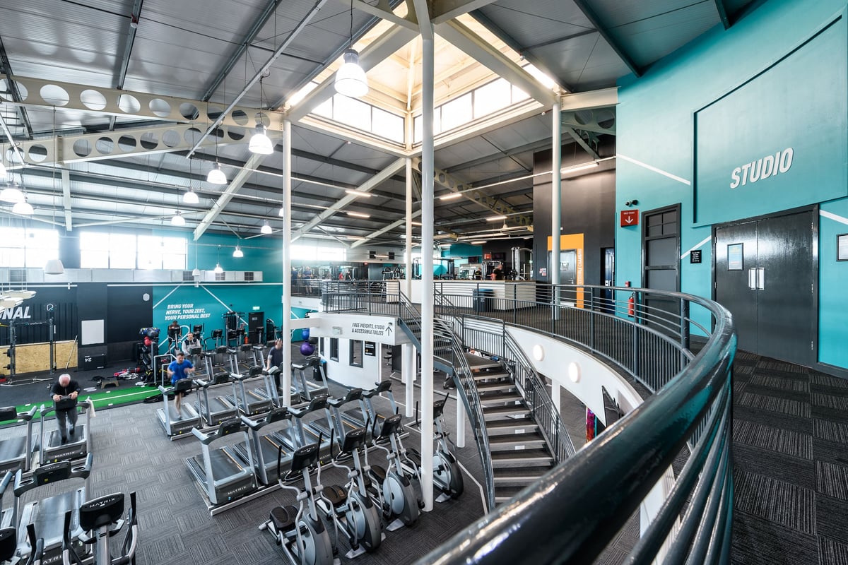 PureGym Manchester Moston: Rows of cardio machines, weight racks, benches, and stairs inside a gym studio.