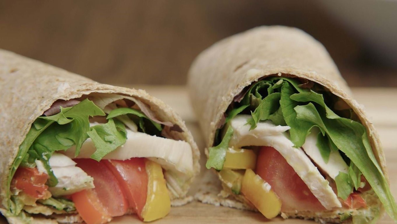 Two halves of a wrap filled with lettuce, tomato, cheese, and yellow pepper on a wooden board