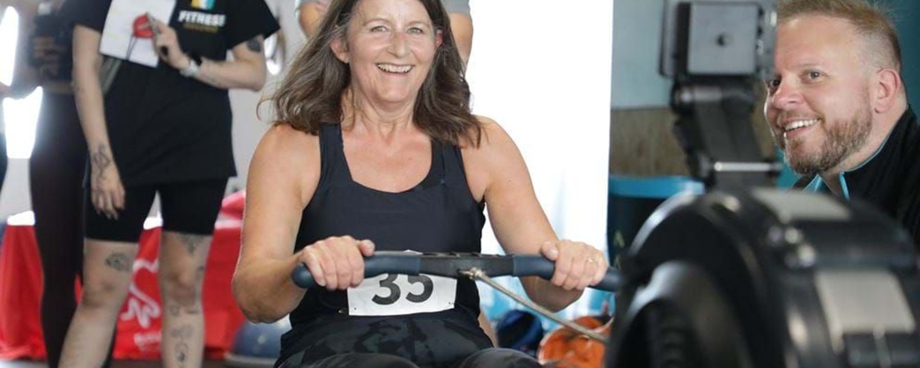 Woman smiling while using a rowing machine with people watching in the background
