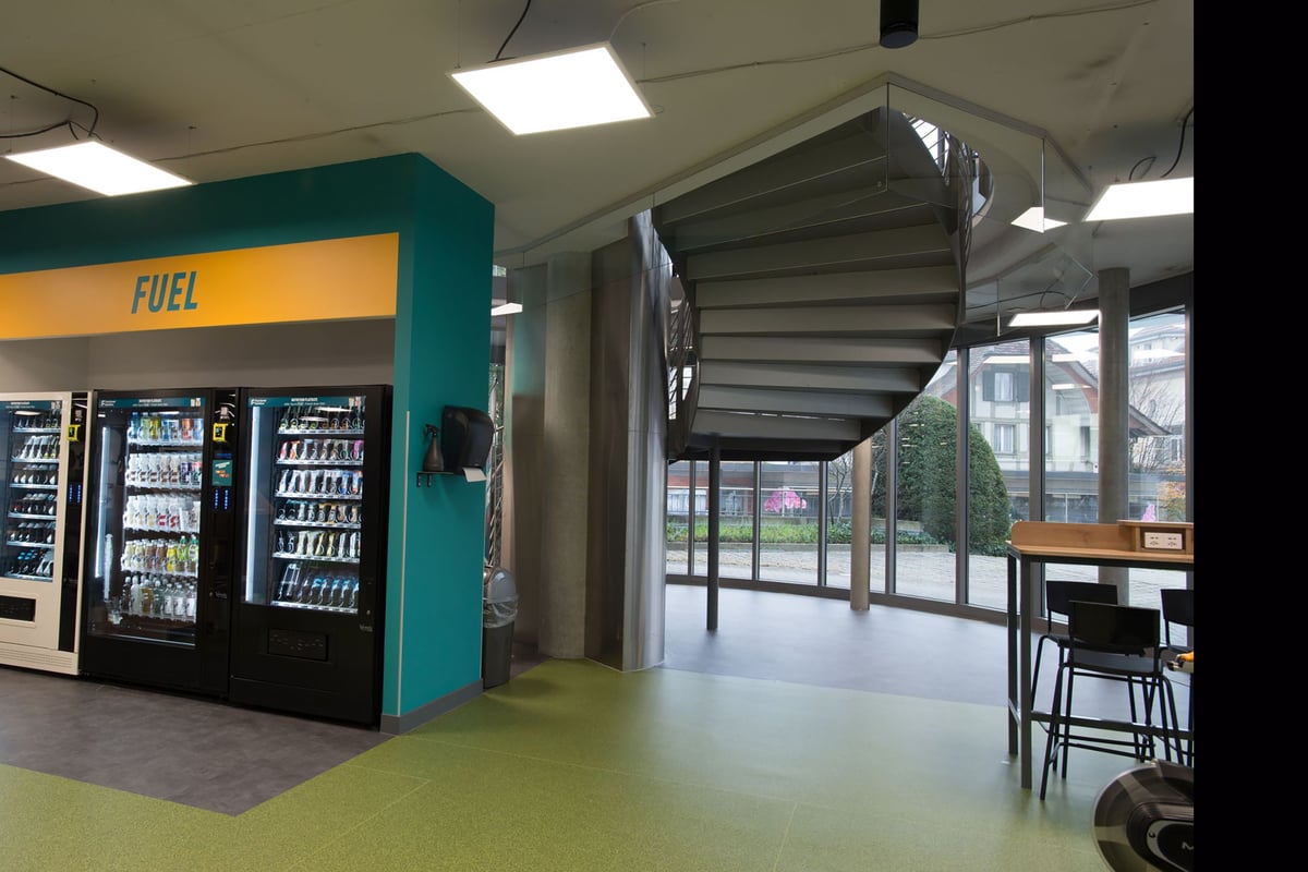 PureGym Lyss: Distributeurs automatiques de carburant, coin salon et portail d'entrée visibles à l'intérieur d'un espace commercial.