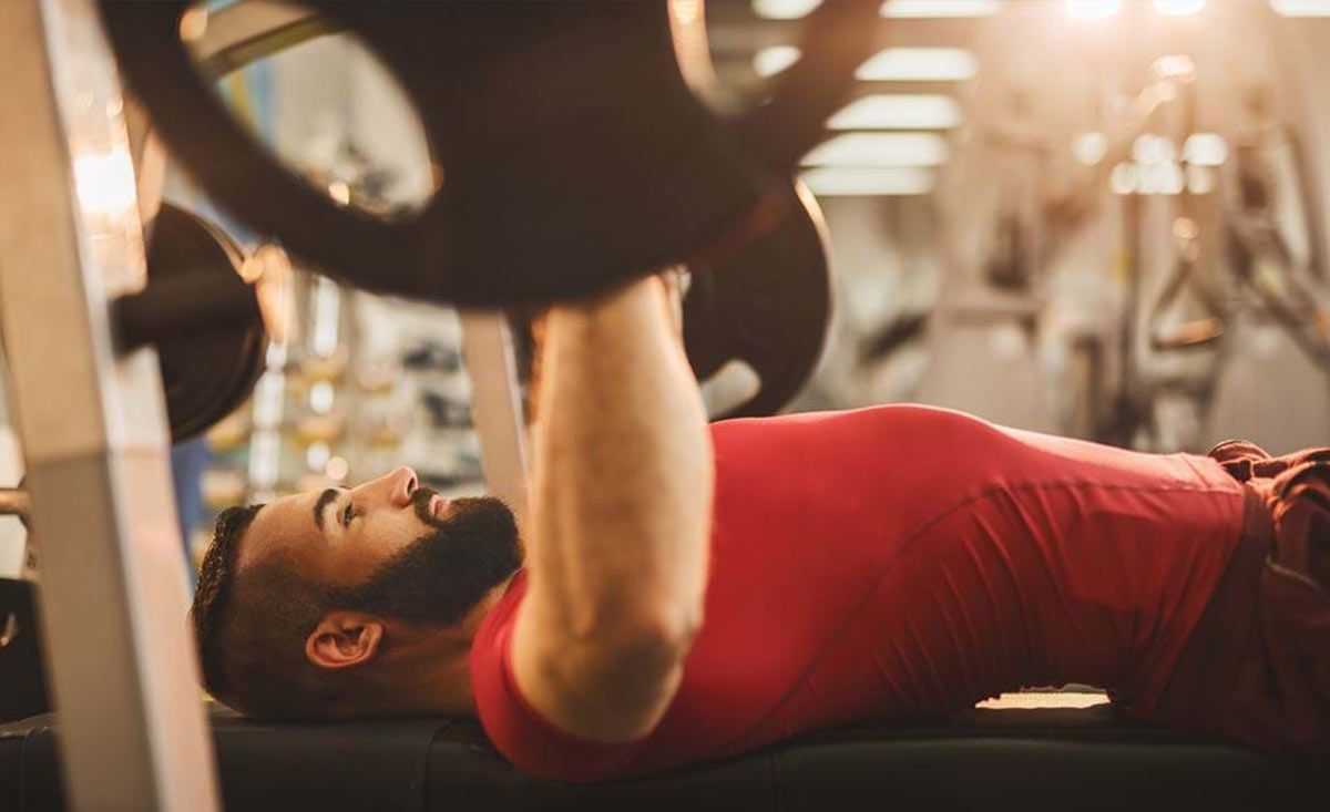 Mann im roten Shirt hebt Gewichte auf der Bank im Fitnessstudio mit Sonnenlichtschein