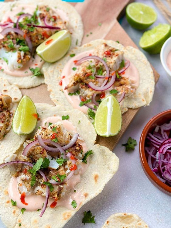 Tortillas topped with grilled fish, red onion, sauce, herbs, and lime wedges on a board