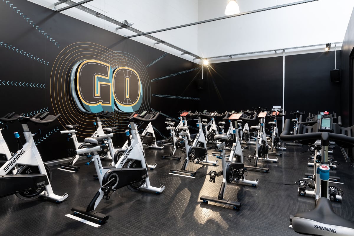 PureGym Colchester Retail Park: Rows of stationary bikes and treadmills in a modern gym with neon GO signage.