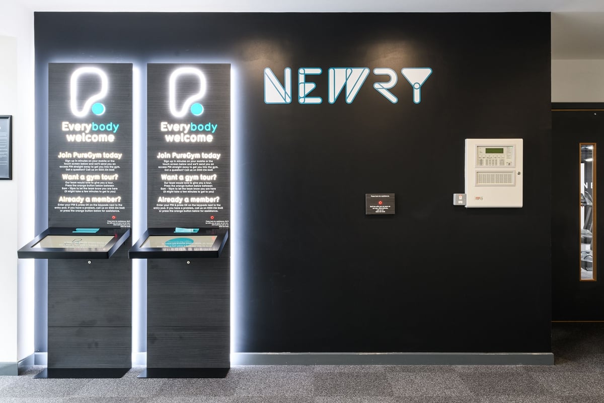 PureGym Newry: Two arcade-style kiosks with screens displaying "Everybody Active" and illuminated "NEWRY" signage.