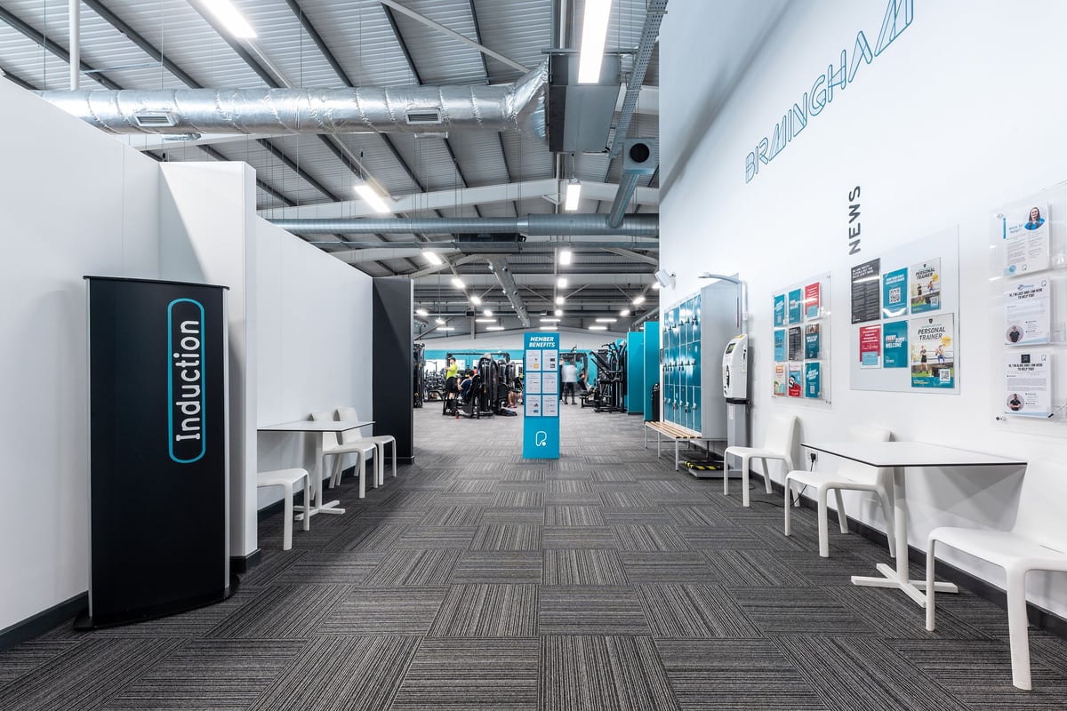 PureGym Birmingham West: Rows of benches, free weights, and lockers with corridor in monochrome tones.