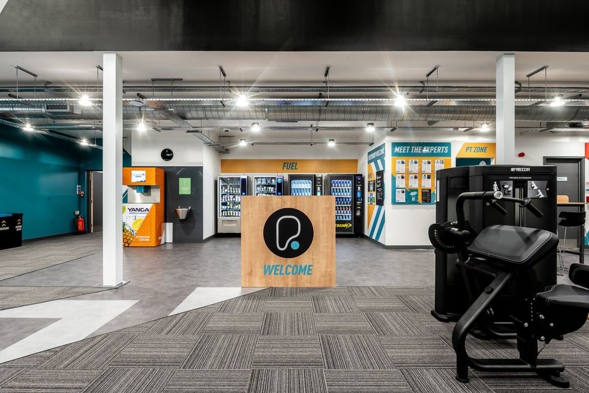 PureGym Ashford Warren Retail Park: Cardio machines, self-service kiosks, lockers, and suspended lighting in a gym facility.