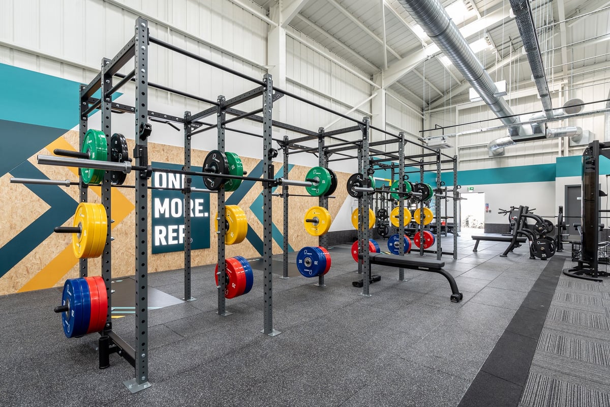 PureGym Keighley: Rigs, weighted plates in varied colours, rubberized floors, rowers and benches visible.