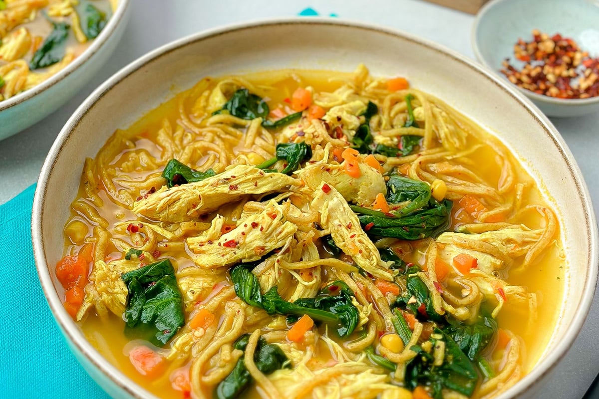 Bowl of chicken noodle soup with yellow broth, spinach, carrots and chilli flakes