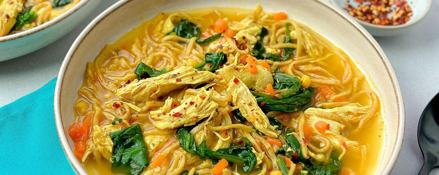 Bowl of chicken noodle soup with yellow broth, spinach, carrots and chilli flakes