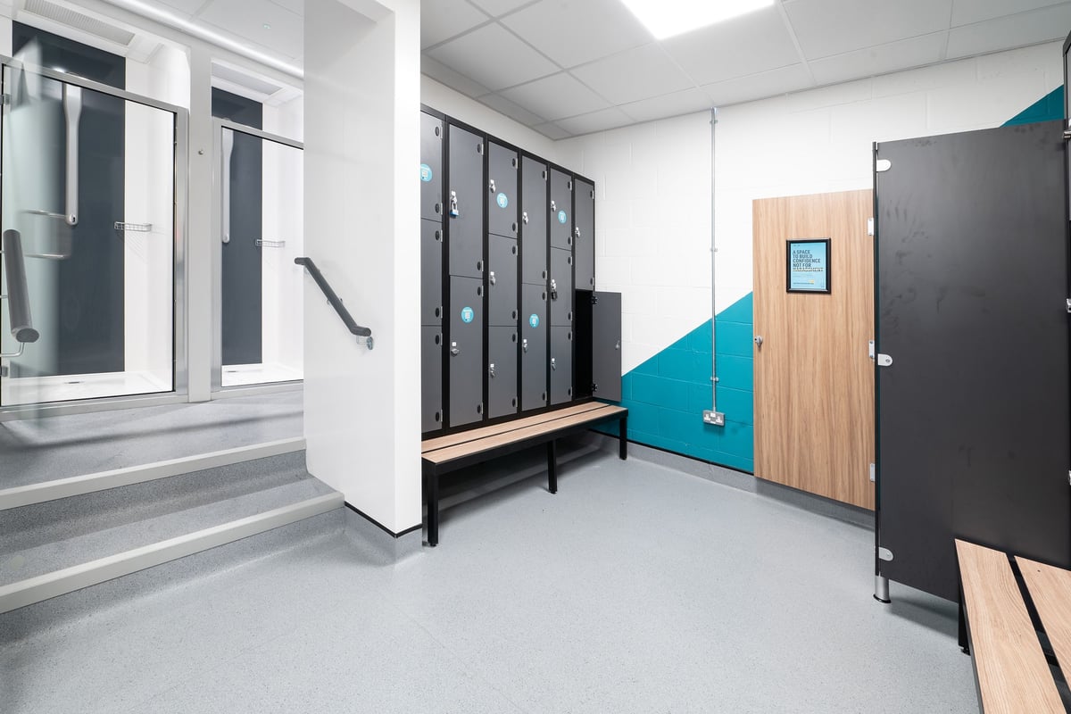 PureGym Dover: Locker area with cubicles, benches, and a digital screen.
