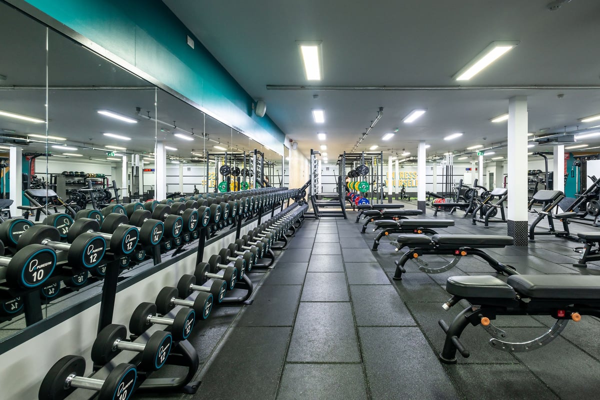 PureGym Wrexham: Rows of exercise bikes, dumbbell racks, weight benches, and rubberised flooring in a bright gym.