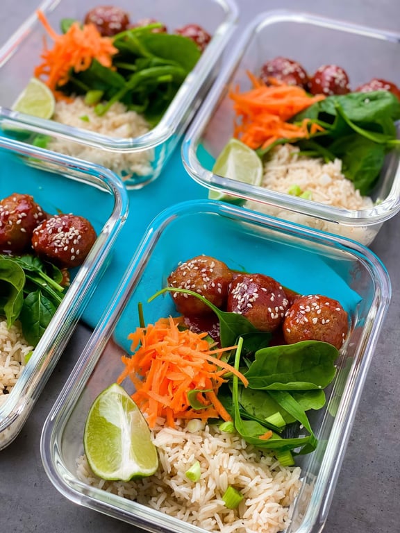 Meal prep containers with rice, shredded carrot, spinach, lime wedge, and meatballs with s