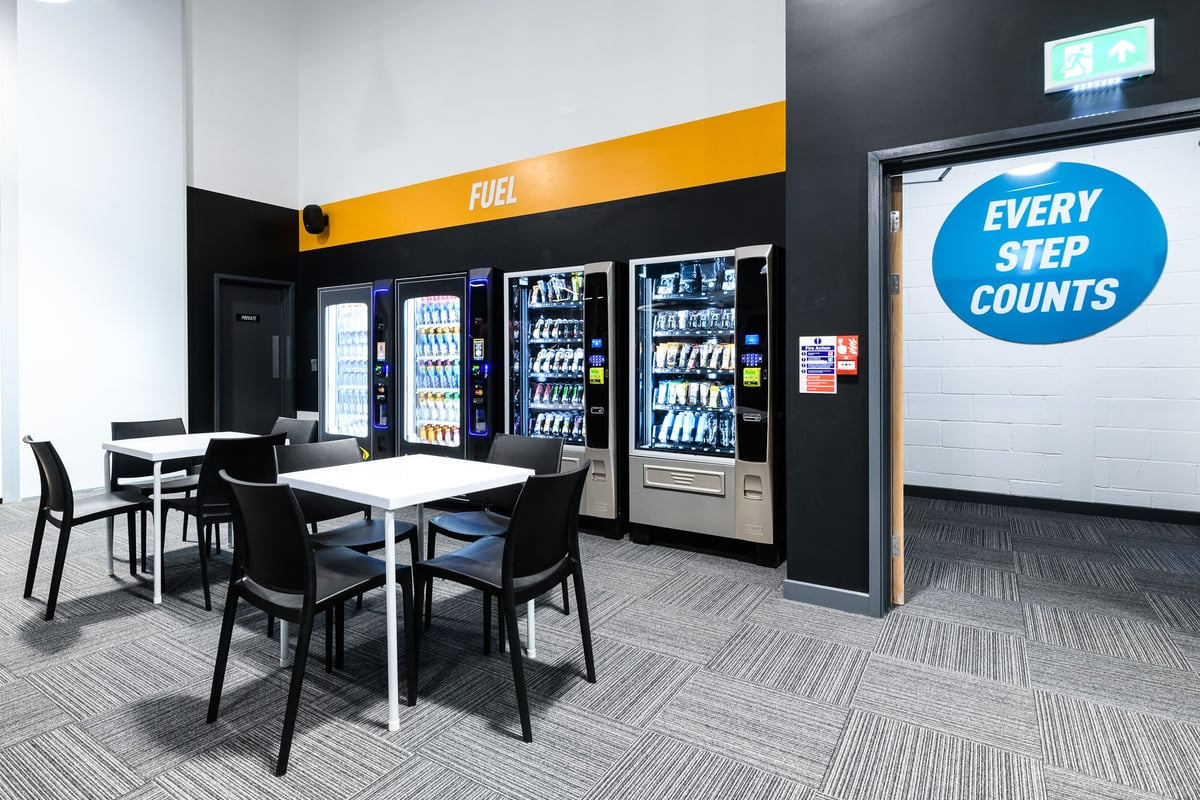 PureGym Cardiff Bay: Vending machines stocked with beverages, table and chairs in a seating area.