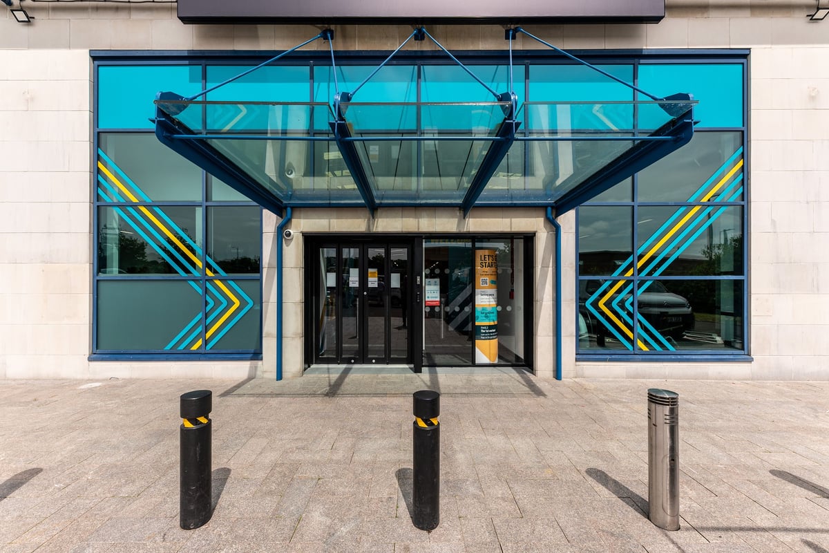 PureGym Coleraine: Glass entrance facade with decorative blue patterns, bollards outside.