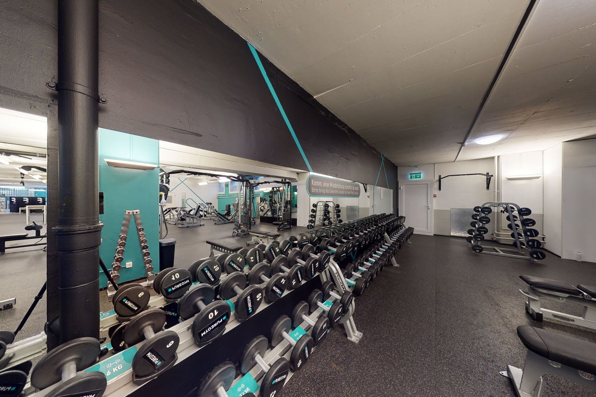 PureGym Basel Dreispitz: Gym studio with free weights, weight machines, and rubberized flooring in teal and grey tones.