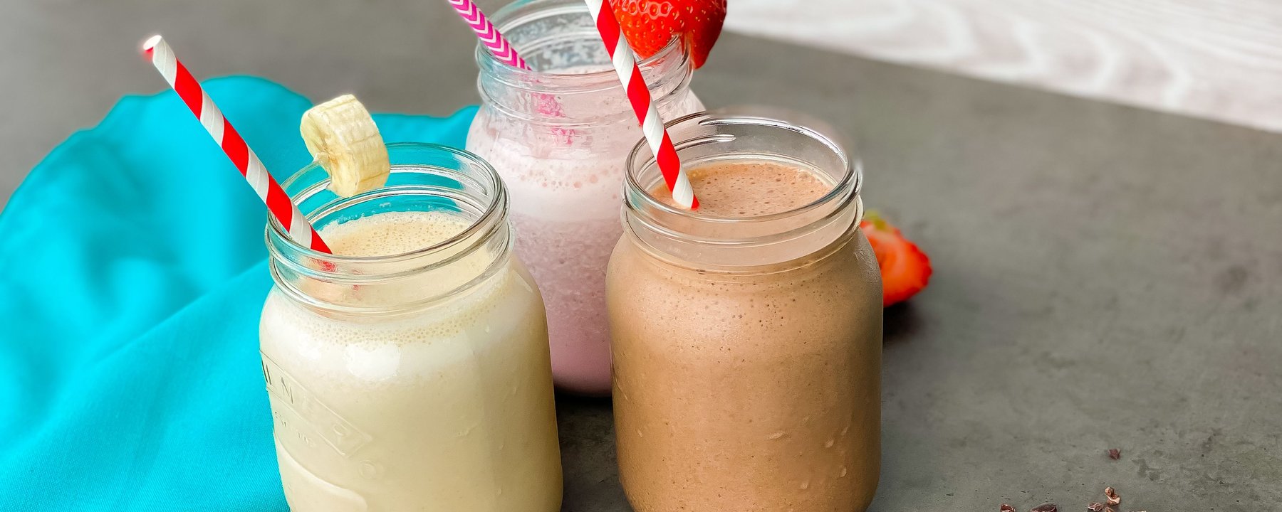 Three protein smoothies in glass jars with striped straws on a grey surface