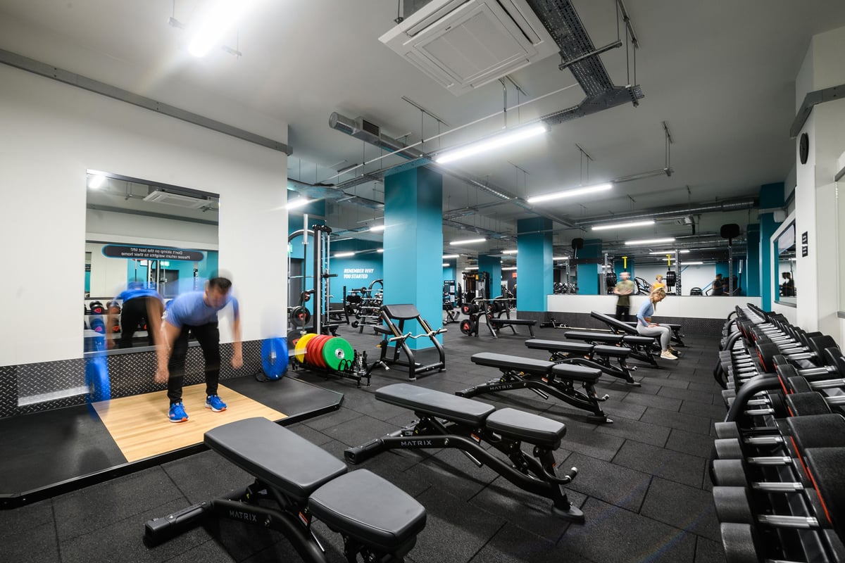 PureGym London Whitechapel: Weights racks, benches, rubberized floors in a teal and grey gym with people exercising.
