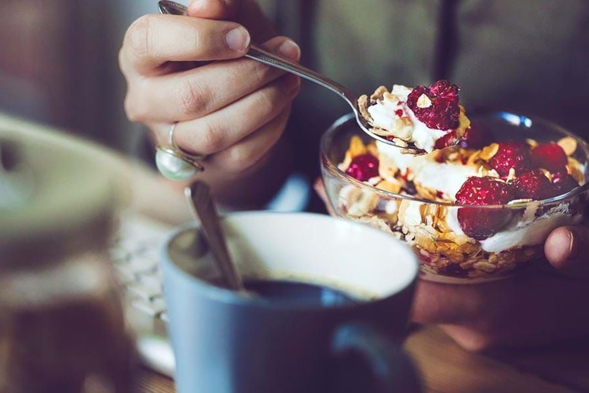 Hand holding spoon with yoghurt, raspberries, and granola above a glass bowl and coffee cu
