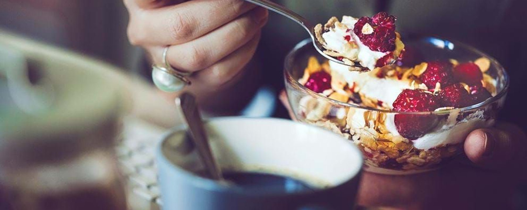 Hand holding spoon with yoghurt, raspberries, and granola above a glass bowl and coffee cu