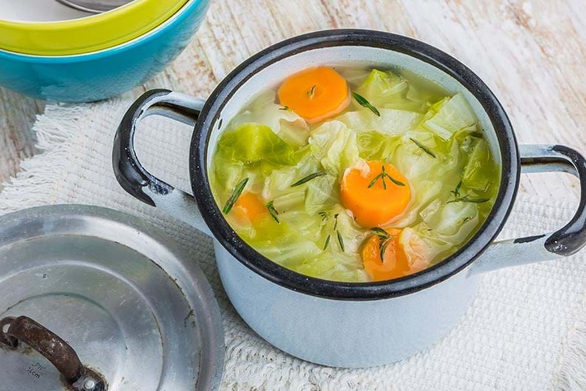 Pot of vegetable soup with carrots, cabbage, and herbs on a white cloth.