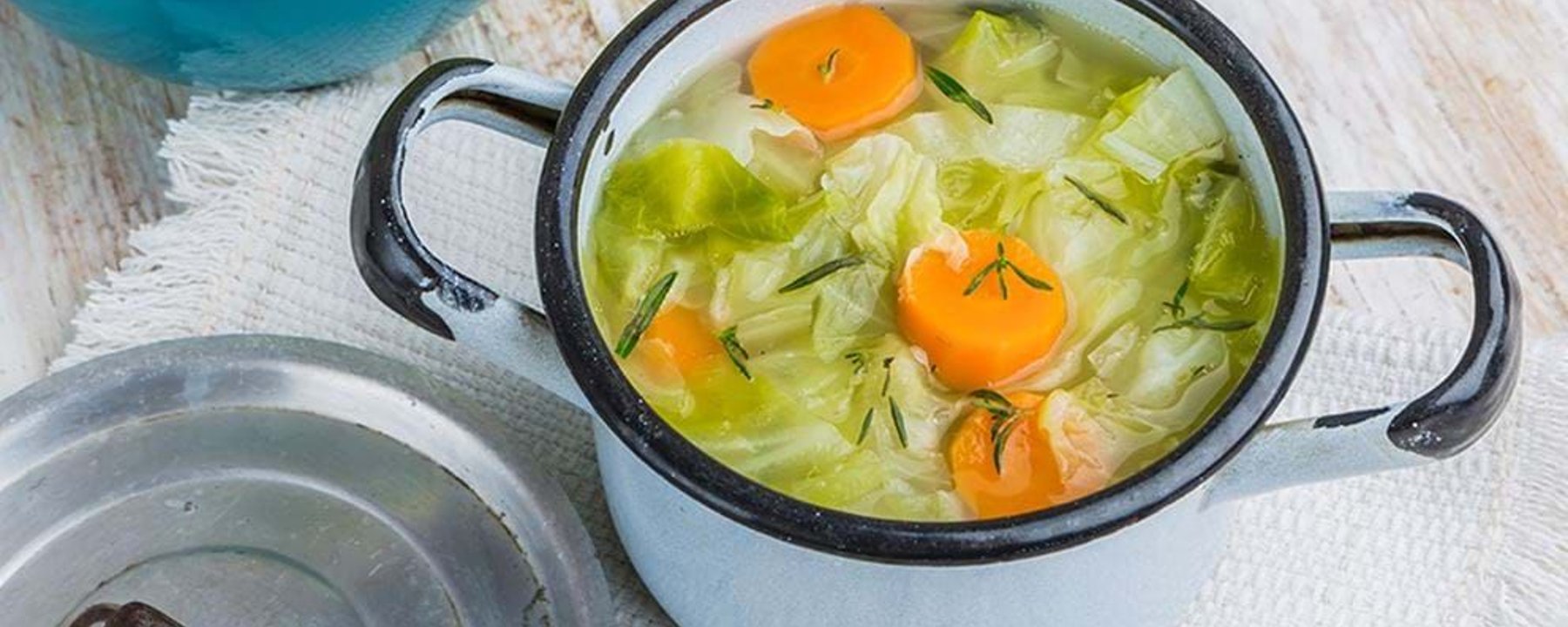 Pot of vegetable soup with carrots, cabbage, and herbs on a white cloth.