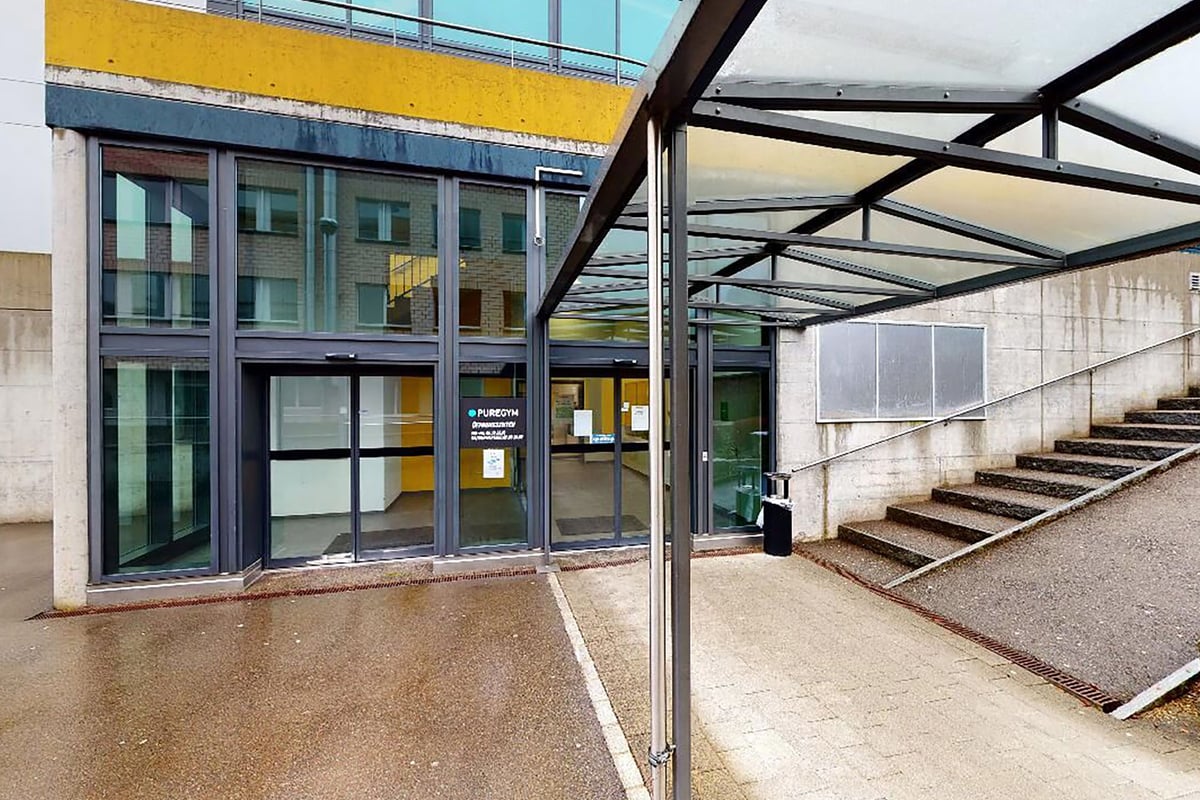 PureGym Pfäffikon: Eingang zum Fitnessstudio mit Glaswänden, Stahlrahmen und Betonböden mit Metalltreppe im Freien.