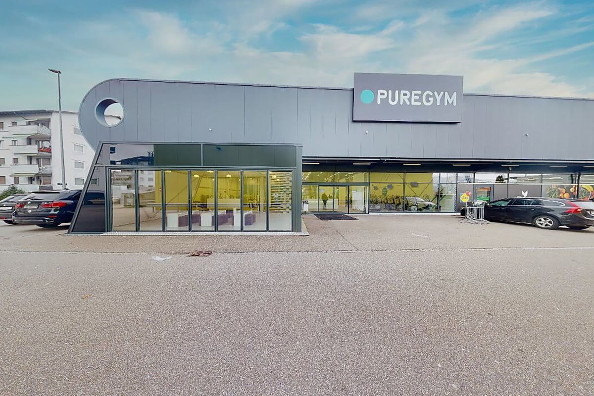 PureGym Oftringen: Exterior of gym franchise with large windows, showing exercise equipment like benches and free weights.