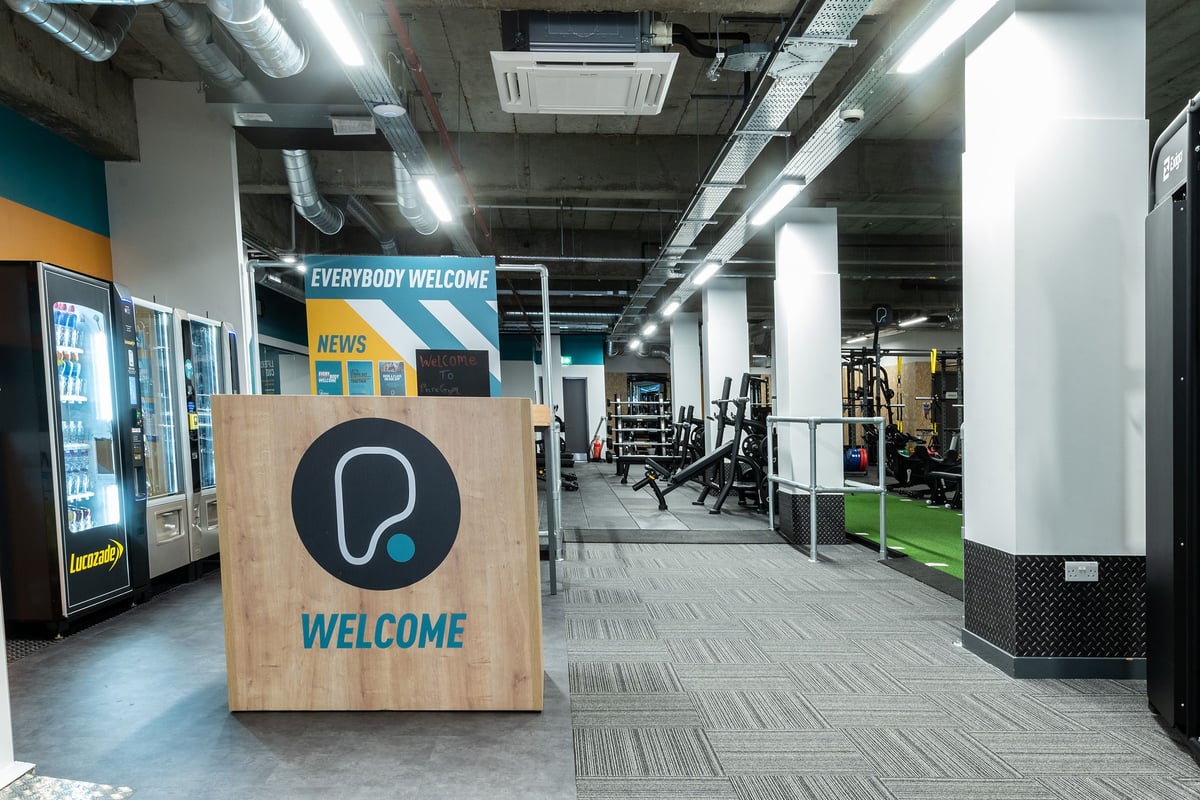 PureGym London Streatham: Welcome sign, free weights area with racks, rubberized flooring, and vending machines.