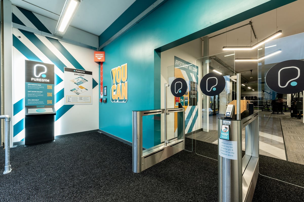 PureGym Solihull Sears Retail Park: Turnstiles, posters, reception desk with check-in kiosks at gym entrance.