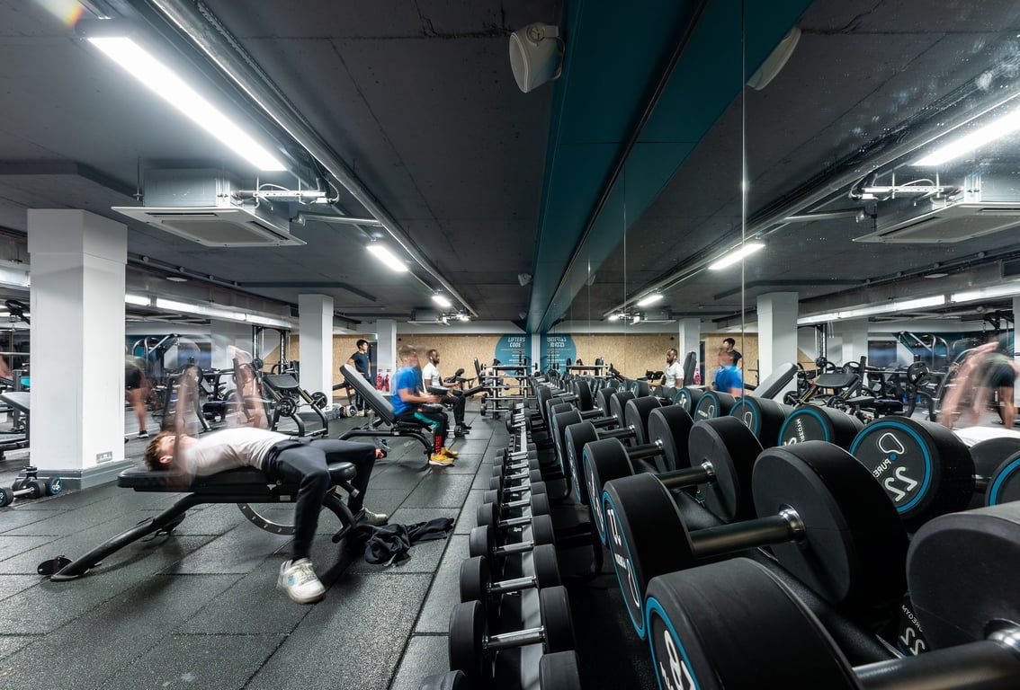PureGym London Putney: Rows of stationary bikes, weight benches, dumbbells, and mirrors in a dimly lit gym interior.