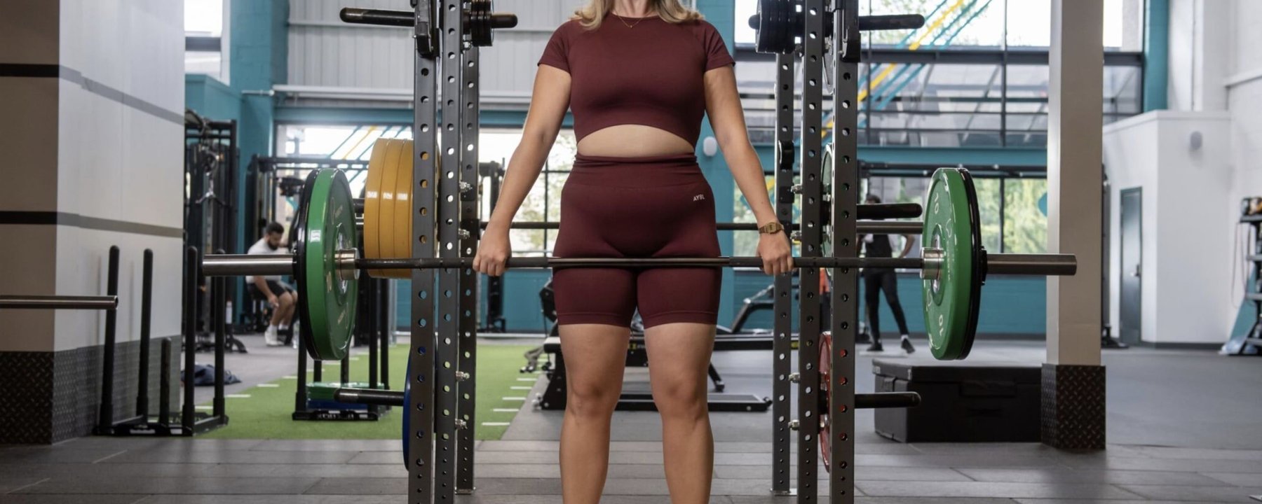 Woman in burgundy sportswear lifting a barbell with green weights in a gym