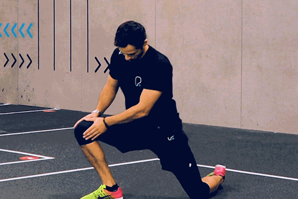 Man in black sportswear performing a kneeling lunge stretch on a gym floor