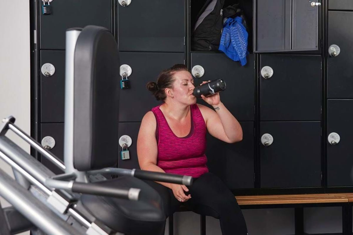 Woman in pink top drinking from a bottle while sitting on a bench in a locker room