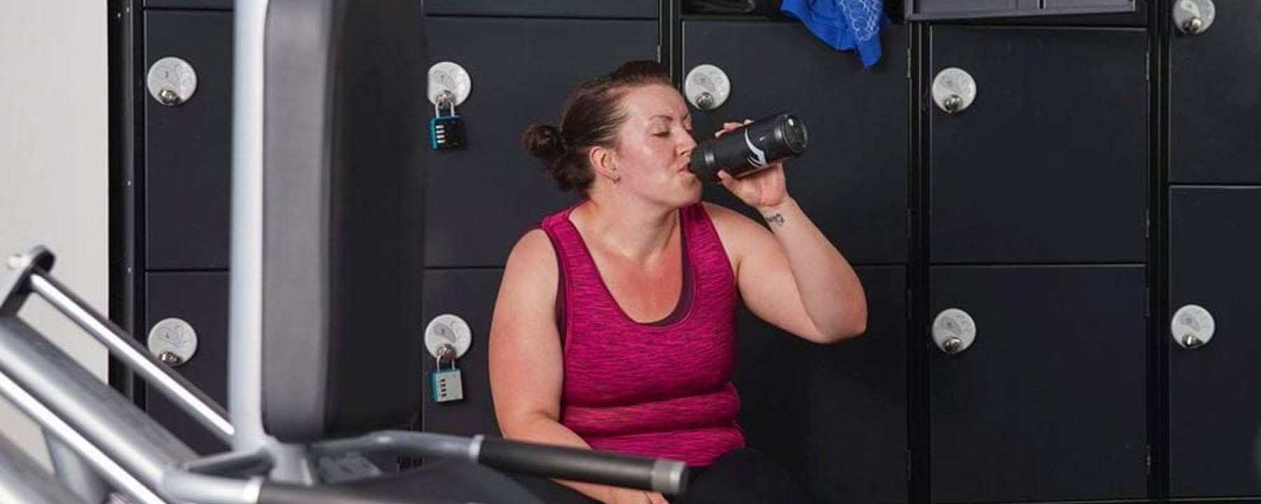 Woman in pink top drinking from a bottle while sitting on a bench in a locker room