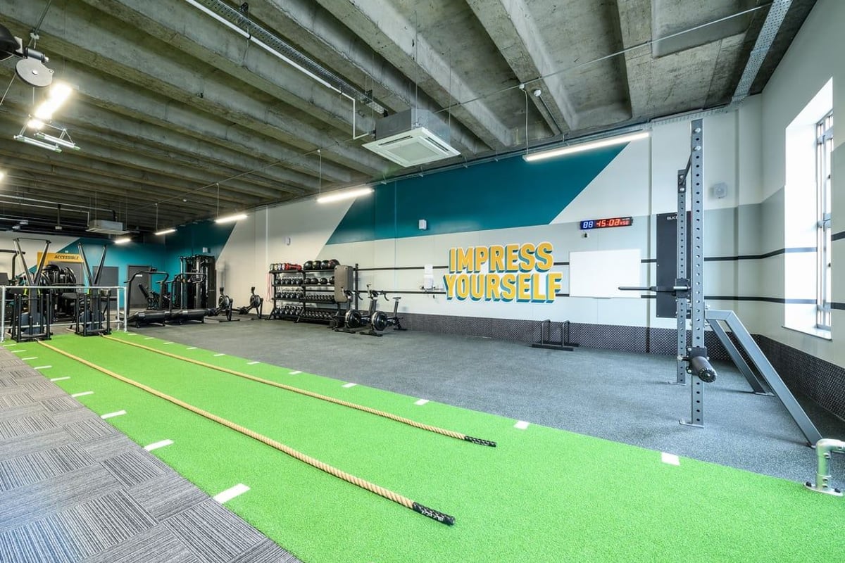 PureGym London Wimbledon: Green turf, free weights, mirrors along wall, exposed ceiling with functional training area.
