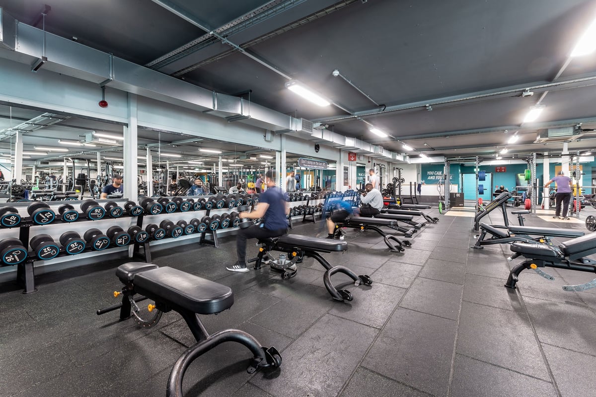 PureGym Oldham: Rows of free weights, benches, resistance machines, and rubberized flooring in a dimly lit gym interior.