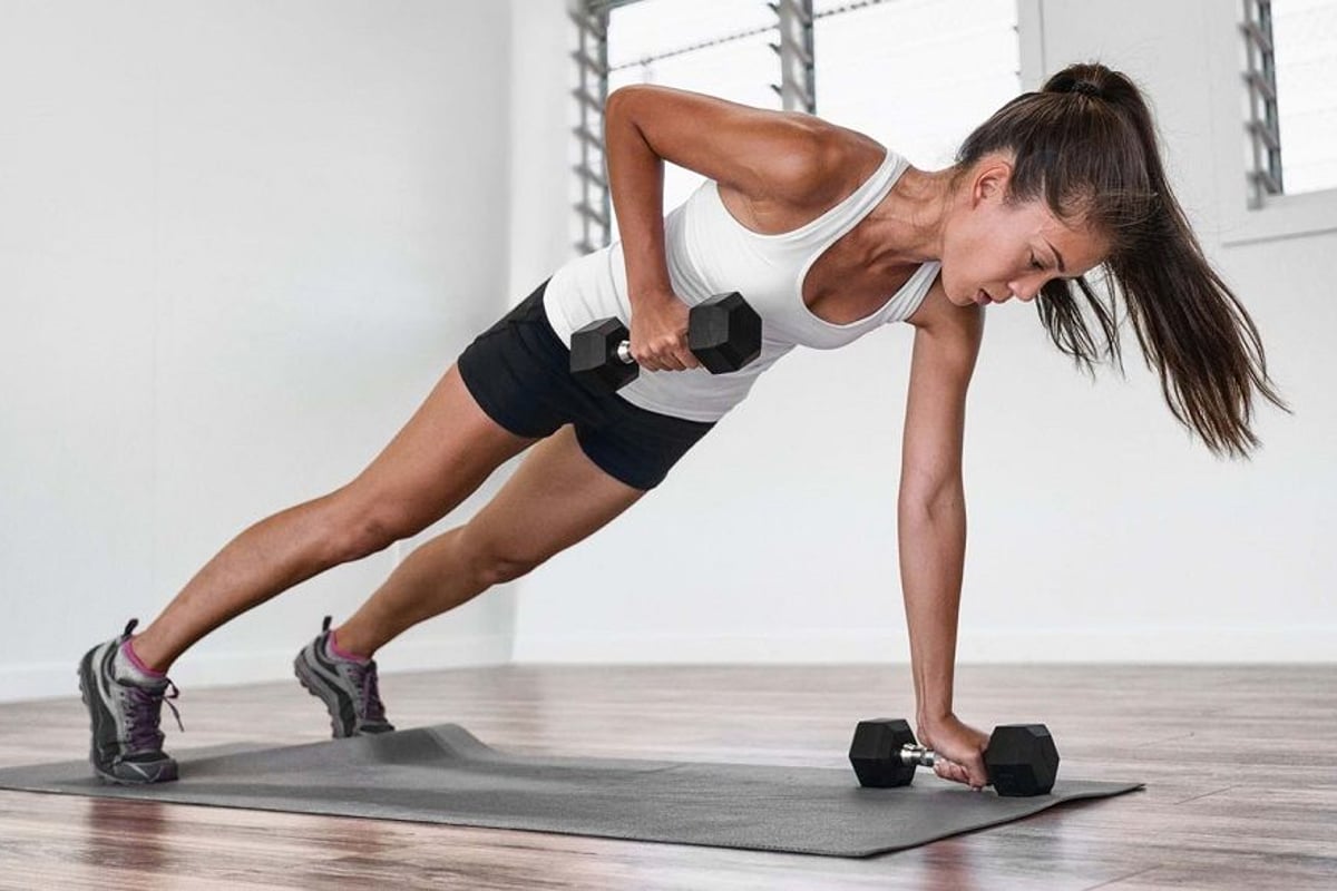 Woman in sportswear doing dumbbell row in plank position on exercise mat indoors
