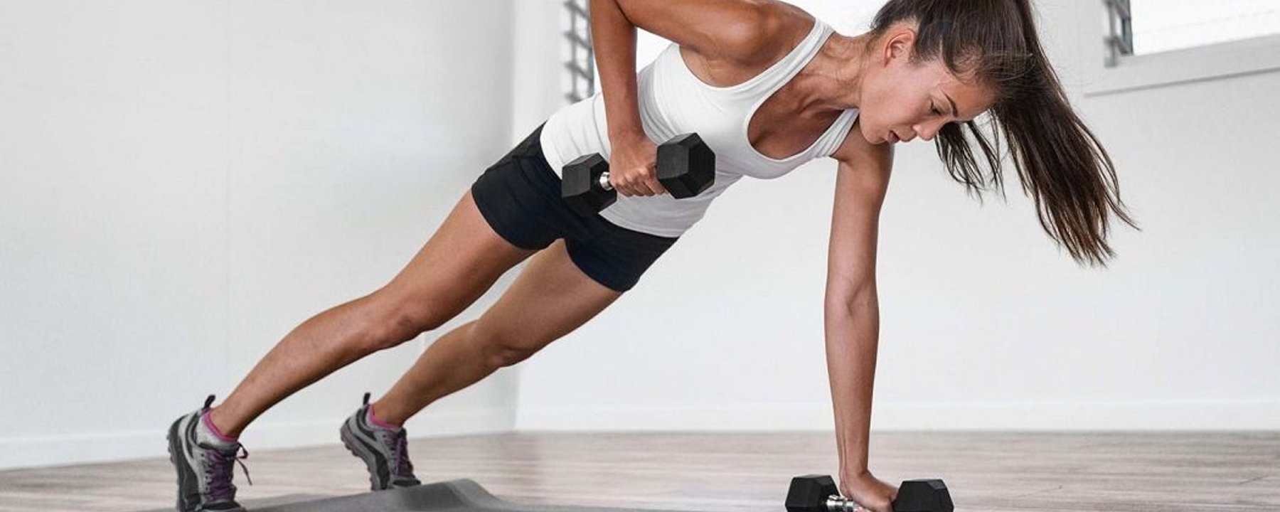 Woman in sportswear doing dumbbell row in plank position on exercise mat indoors