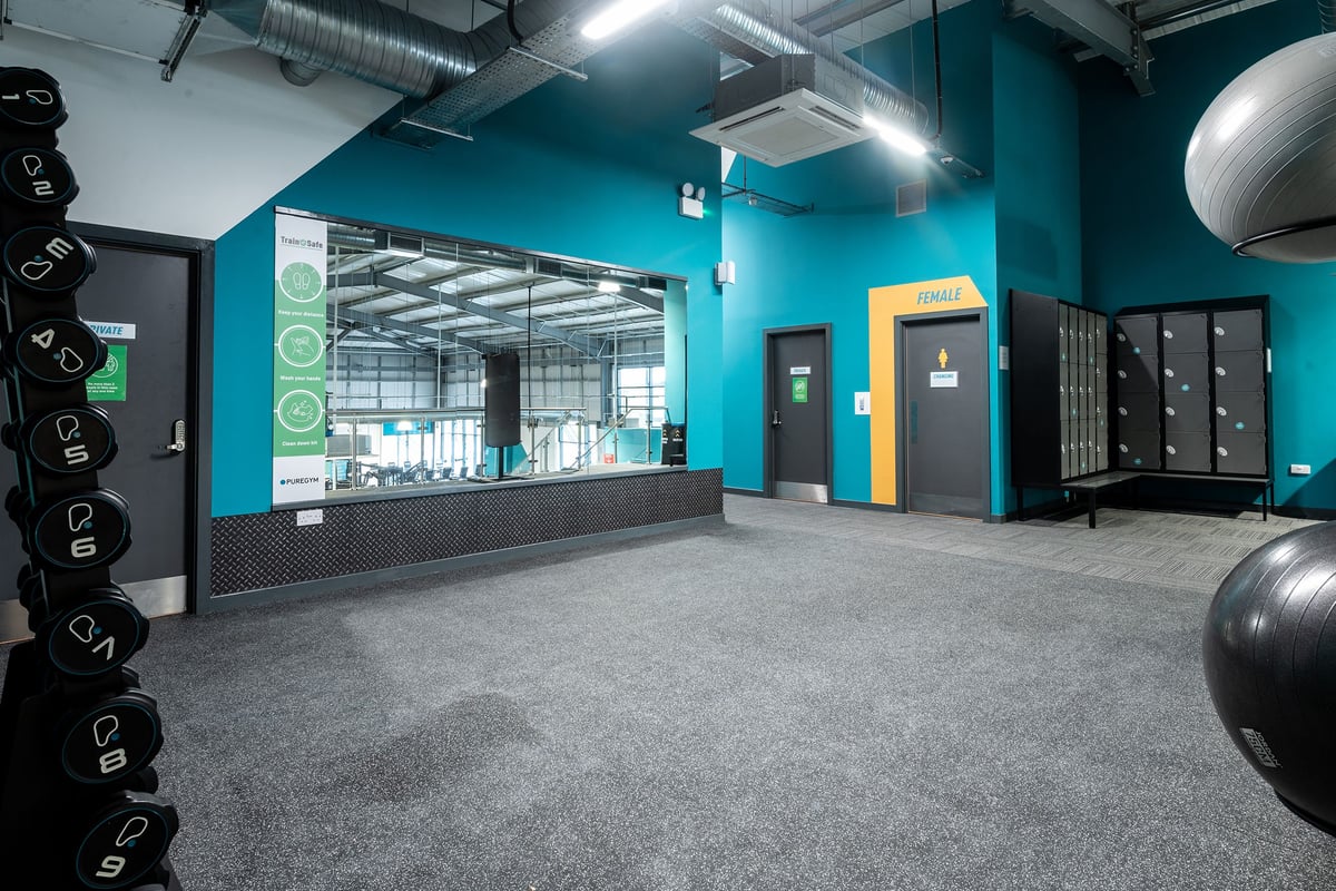 PureGym Cirencester: Weight balls, lockers, reception desk, teal and yellow accent walls in fitness studio.