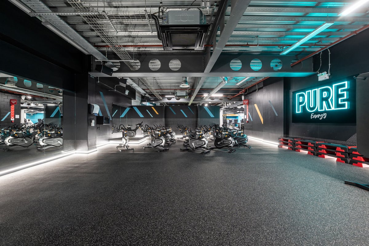 PureGym Manchester Spinningfields: Rows of stationary bikes, neon "PURE" signage, industrial ceiling, rubberized flooring.