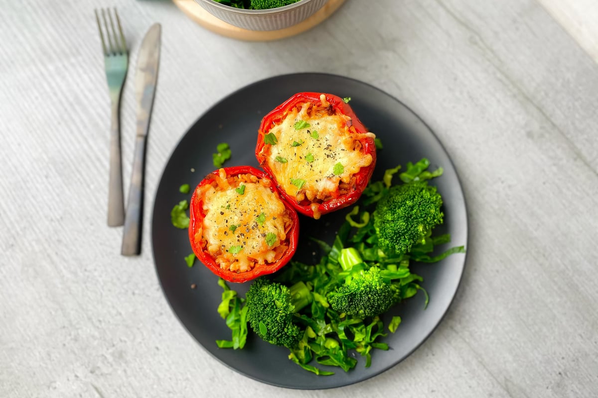 Two stuffed red peppers with melted cheese and herbs on a black plate with broccoli