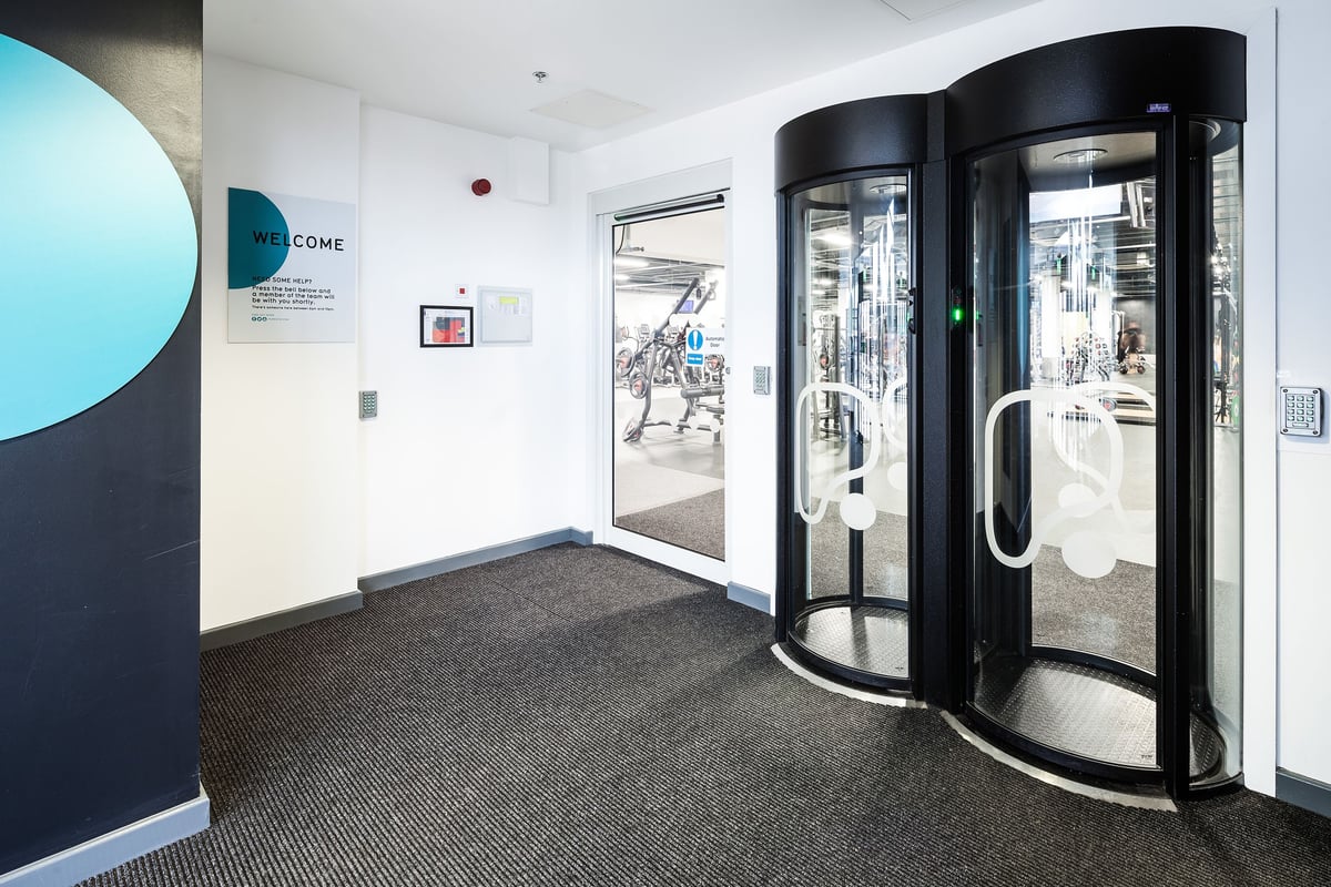 PureGym Altrincham: Revolving doors, reception area with teal signage and carpet flooring.
